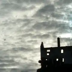 A dark silhouette of a lighthouse and attached building stands against a cloudy sky.