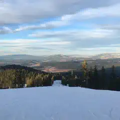 A snow-covered slope descends toward a valley with distant mountains and evergreen trees.
