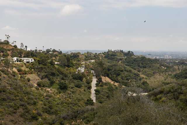 A paved road goes down a tree-covered hillside. Houses are on the slopes, and a city is in the distance.