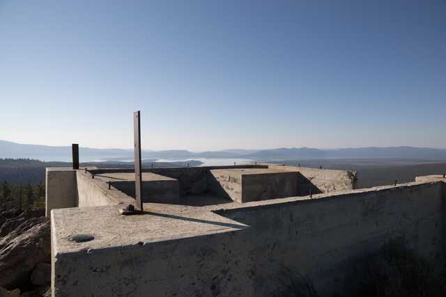 A concrete structure stands on a rocky outcrop overlooking a lake and distant mountains under a clear blue sky.