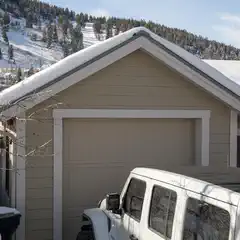 A tan garage with a snow-covered roof is adjacent to a white Jeep and a hillside covered in evergreen trees.