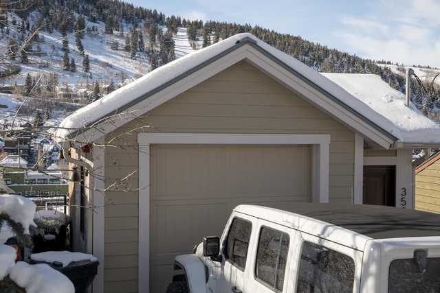 A tan garage with a snow-covered roof is adjacent to a white Jeep and a hillside covered in evergreen trees.