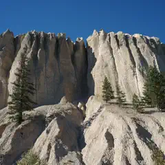 A formation of tan rock spires rises from a hillside with scattered evergreen trees under a clear blue sky.