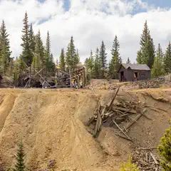 An abandoned wooden structure and debris sit on a dirt hillside, flanked by two small log cabins and a forest of pine trees.