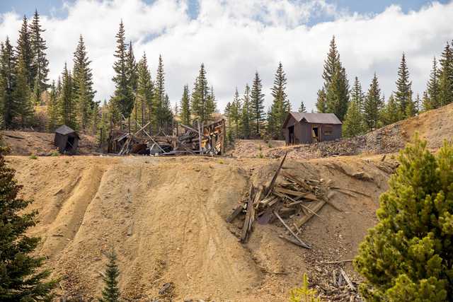 An abandoned wooden structure and debris sit on a dirt hillside, flanked by two small log cabins and a forest of pine trees.