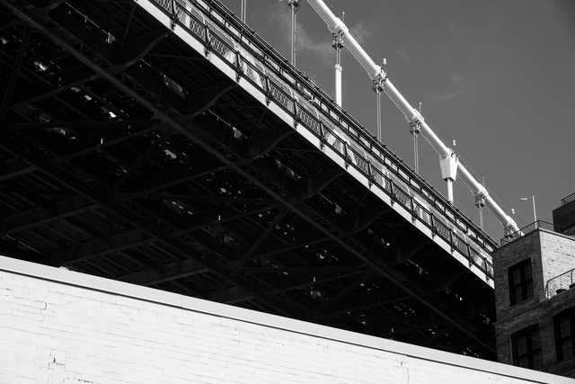 A bridge's intricate steel framework and ornate railing are captured from below, with a brick building in the foreground.