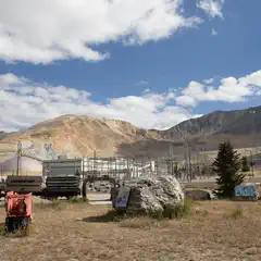 A field of dry grass fronts heavy mining equipment and industrial buildings against a backdrop of mountain slopes.