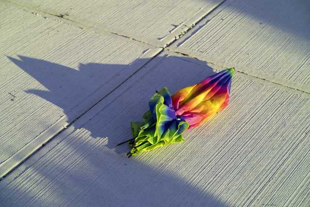 A colorful, collapsed umbrella lies on a textured concrete surface, casting a sharp shadow in bright sunlight.