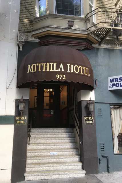 A brown awning with gold lettering marks the entrance to a hotel, situated above a set of stairs leading up to the building's main door.