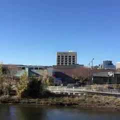A multi-story building stands among lower, adjacent structures, with a body of water and trees in the foreground.