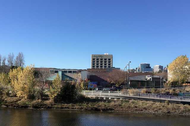 A multi-story building stands among lower, adjacent structures, with a body of water and trees in the foreground.