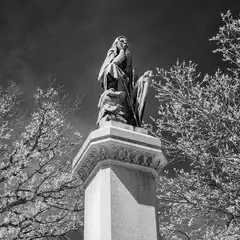 A statue of a draped figure stands atop a substantial stone pillar, flanked by trees.