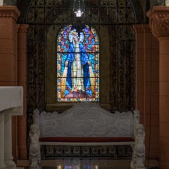 A stained glass window depicts a robed figure with a halo, surrounded by blue and red hues. A bench with ornate carvings sits below the window.