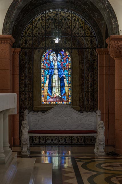 A stained glass window depicts a robed figure with a halo, surrounded by blue and red hues. A bench with ornate carvings sits below the window.