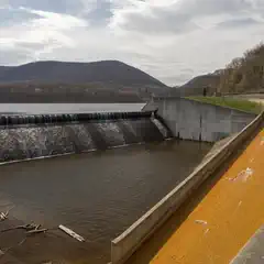A concrete dam and spillway, surrounded by forested hills and a yellow-tinted waterway.