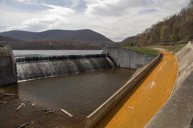 A concrete dam and spillway, surrounded by forested hills and a yellow-tinted waterway.