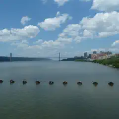 A suspension bridge spans a wide body of water with pilings visible in the foreground and buildings and trees lining the shore.