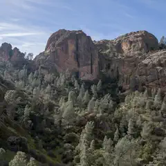 A rugged landscape features towering rock formations and dense coniferous forest under a clear blue sky.