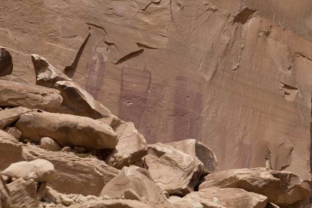 Faded red pictographs of human-like figures are visible on a weathered sandstone wall above a pile of large, angular boulders.