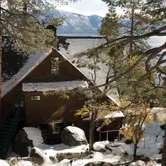 A snow-covered wooden cabin stands among trees with a lake and mountains in the background.