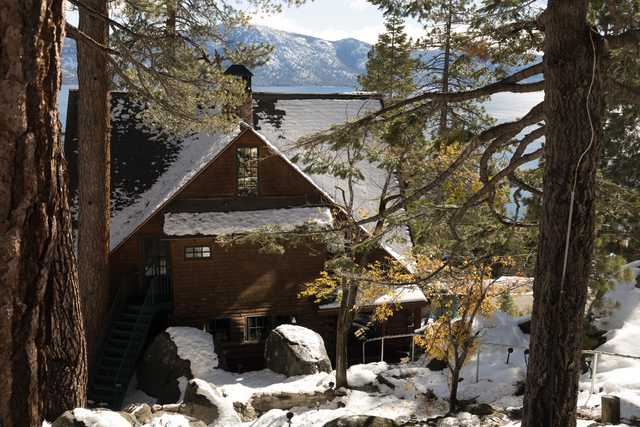 A snow-covered wooden cabin stands among trees with a lake and mountains in the background.