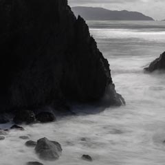 A turbulent ocean flows around dark rocks and cliffs, with an archway in the foreground.