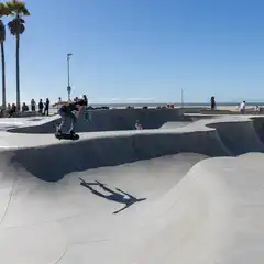 A skateboarder performs a trick in a concrete skate park near a beach, with palm trees and onlookers in the background.