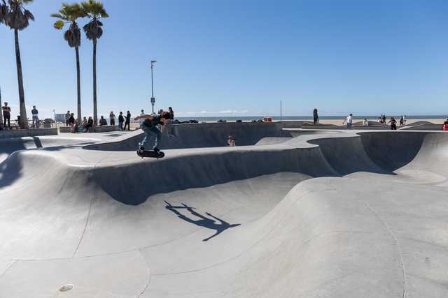 A skateboarder performs a trick in a concrete skate park near a beach, with palm trees and onlookers in the background.