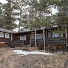 A weathered wooden cabin with a sloped roof, surrounded by trees and rocks, with patches of snow on the ground.