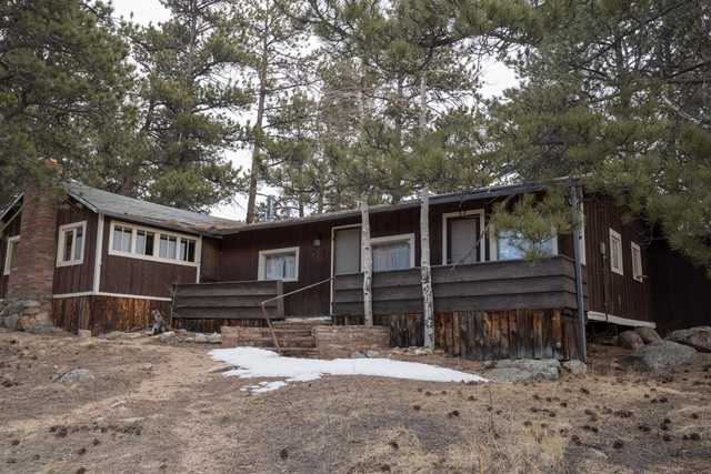 A weathered wooden cabin with a sloped roof, surrounded by trees and rocks, with patches of snow on the ground.
