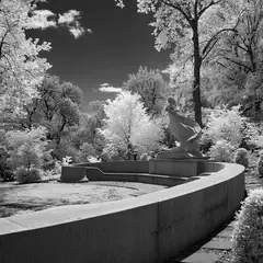 A curved concrete wall borders a landscaped area with a sculpture and trees under a cloudy sky.