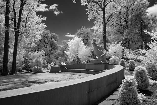 A curved concrete wall borders a landscaped area with a sculpture and trees under a cloudy sky.