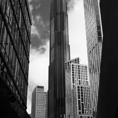 A towering skyscraper dominates a dense urban skyline, framed by surrounding high-rise buildings and a cloudy sky.
