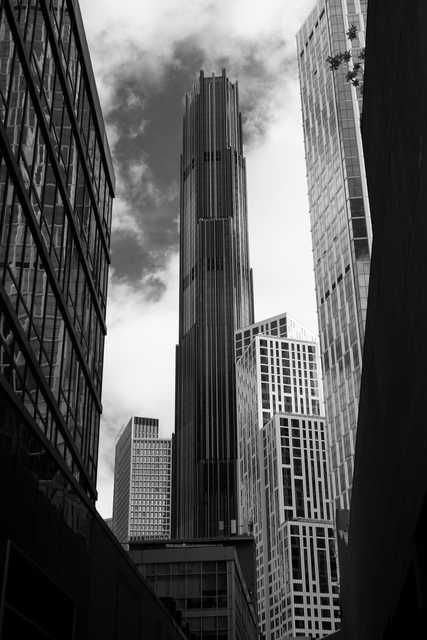 A towering skyscraper dominates a dense urban skyline, framed by surrounding high-rise buildings and a cloudy sky.