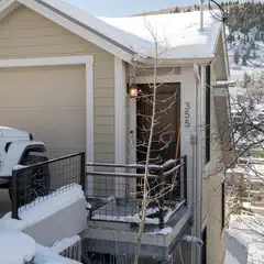A tan house with a snow-covered roof and a balcony railing covered in snow, situated on a hillside with mountains visible in the background.