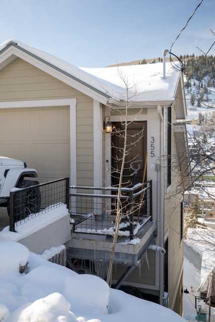 A tan house with a snow-covered roof and a balcony railing covered in snow, situated on a hillside with mountains visible in the background.