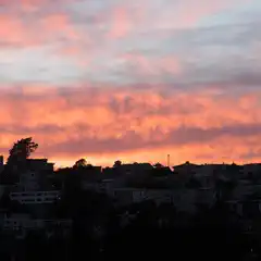 A sunset over a city, with clouds glowing orange and pink against a dark blue sky, and silhouettes of houses on a hillside below.