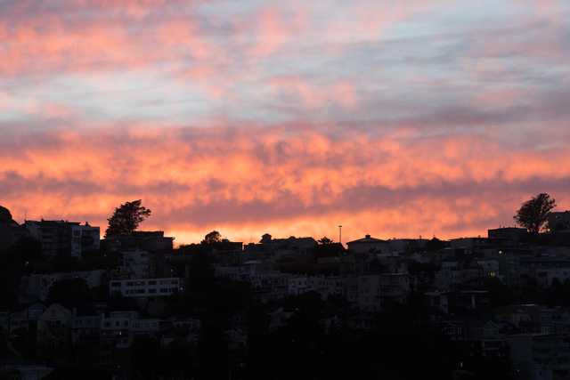 A sunset over a city, with clouds glowing orange and pink against a dark blue sky, and silhouettes of houses on a hillside below.