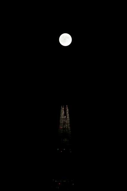 A full moon hangs above the illuminated spire of a tall building at night.