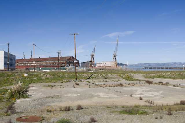 A low building with a rusted corrugated metal exterior sits beside two cranes and a grassy field under a clear blue sky.