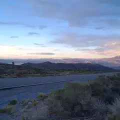 A paved road extends through a desert landscape toward distant mountains under a pink and blue sky.