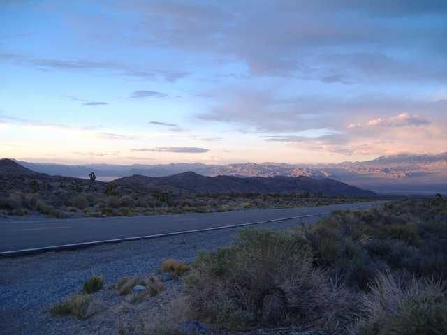 A paved road extends through a desert landscape toward distant mountains under a pink and blue sky.