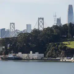 A US Coast Guard vessel sits docked near low, white buildings adjacent to a green hillside with a city skyline and bridge in the background.