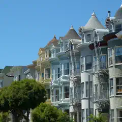 A row of Victorian houses with pointed roofs and bay windows, painted in pastel colors, under a clear blue sky.