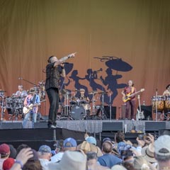 A band performing on stage, with lead singer gesturing to audience and musicians playing instruments behind him.