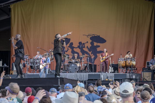A band performing on stage, with lead singer gesturing to audience and musicians playing instruments behind him.
