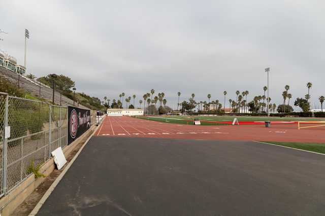 A red running track borders a green football field, with palm trees lining the background and bleachers to the left.