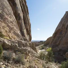 A narrow canyon contains a sandy wash between steep, reddish rock walls under a blue sky.