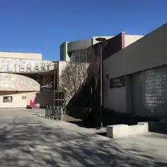 A library building with a curved facade and large windows, featuring a sign that reads "Library". The entrance is visible with steps leading up to it.