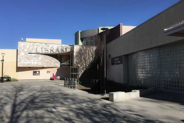 A library building with a curved facade and large windows, featuring a sign that reads "Library". The entrance is visible with steps leading up to it.